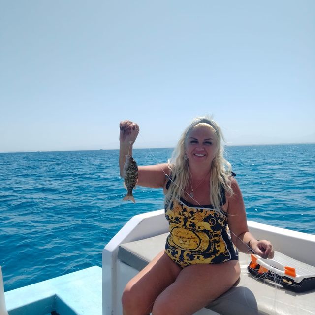 Smiling guest on a boat deck holding up a small fish caught on a fishing line, with open sea behind
