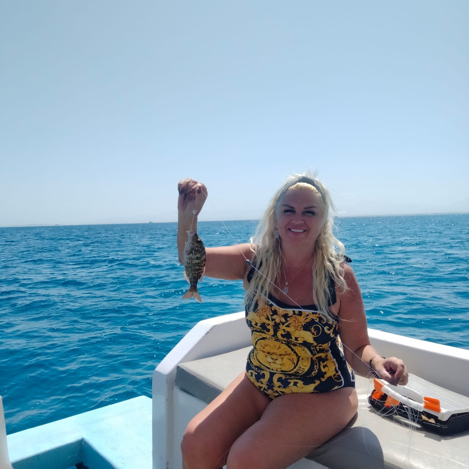 ضيف مبتسم على سطح قارب وهو يرفع سمكة صغيرة اصطادها بخيط الصيد، ويظهر البحر المفتوح خلفه