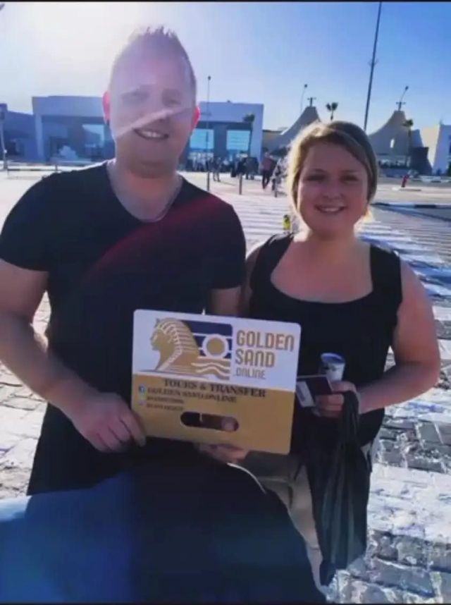 Usmievavý pár s tabuľkou Golden Sand Tours and Transfer pred letiskom v Hurghade