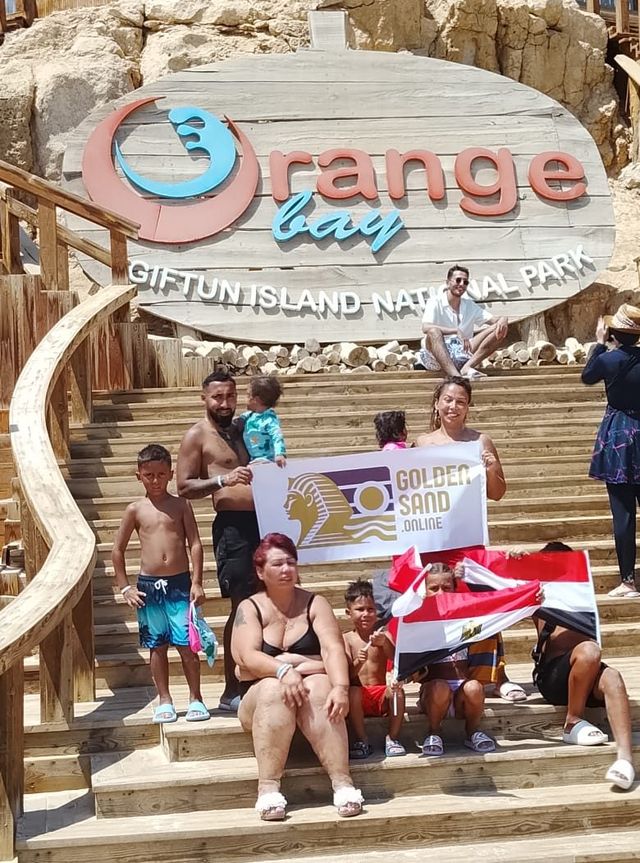 Families with children on the steps below the Orange Bay sign, holding a Golden Sand banner and Egyptian flags