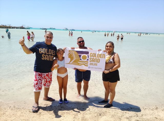 Group of four holding a Golden Sand banner in shallow turquoise water, with boats and a wooden pier behind them