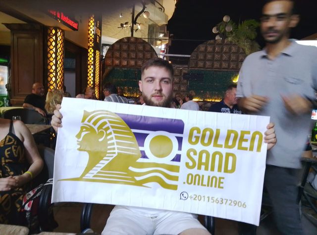 Guest at a cafe table at night holding up the Golden Sand banner as a man in a grey polo walks past