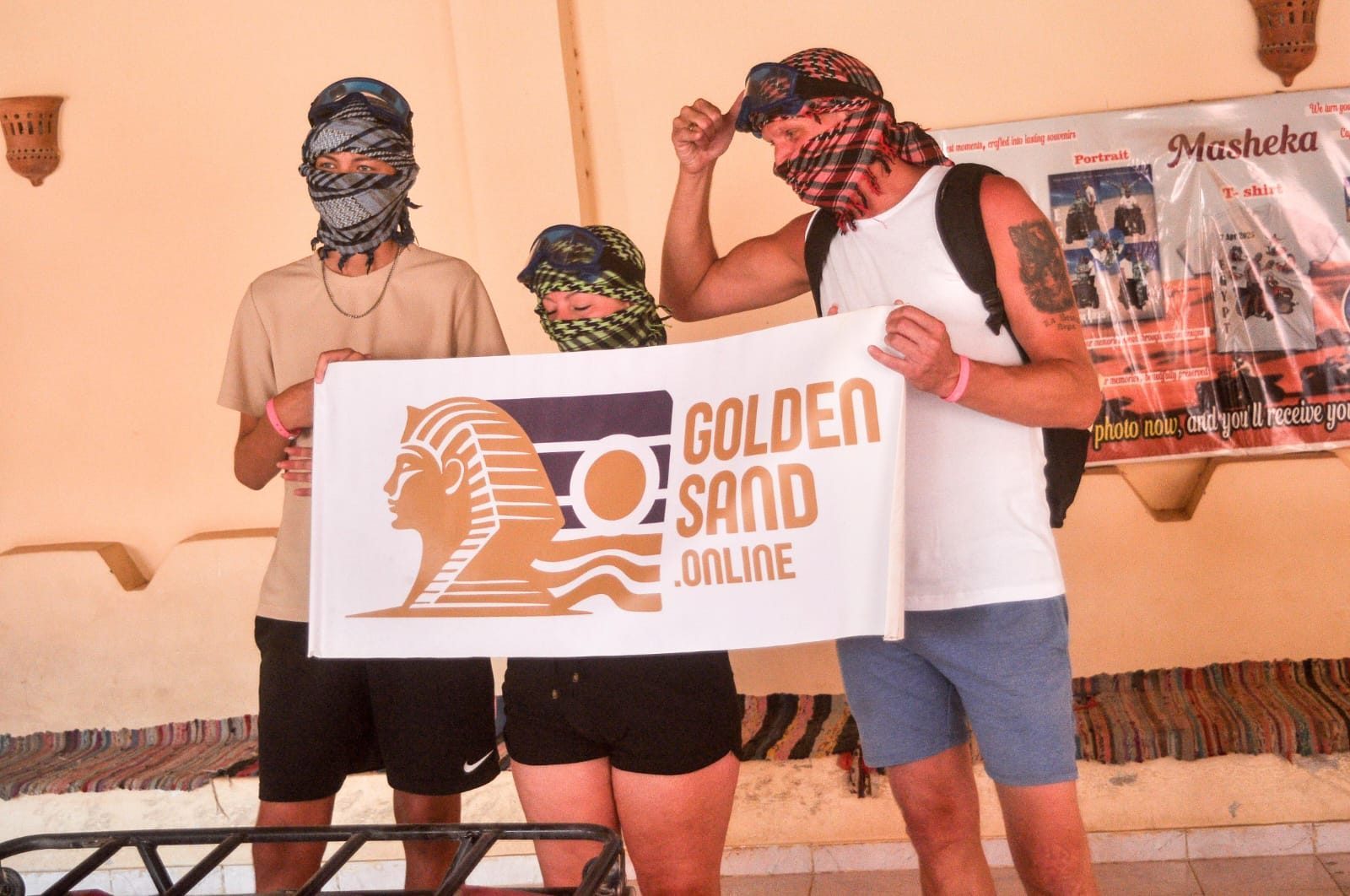 ثلاثة ضيوف يرتدون أوشحة على الوجه ونظارات واقية، يحملون لافتة كبيرة مكتوب عليها «Golden Sand» خلال توقف في رحلة سفاري بالسيارات الرباعية في الصحراء