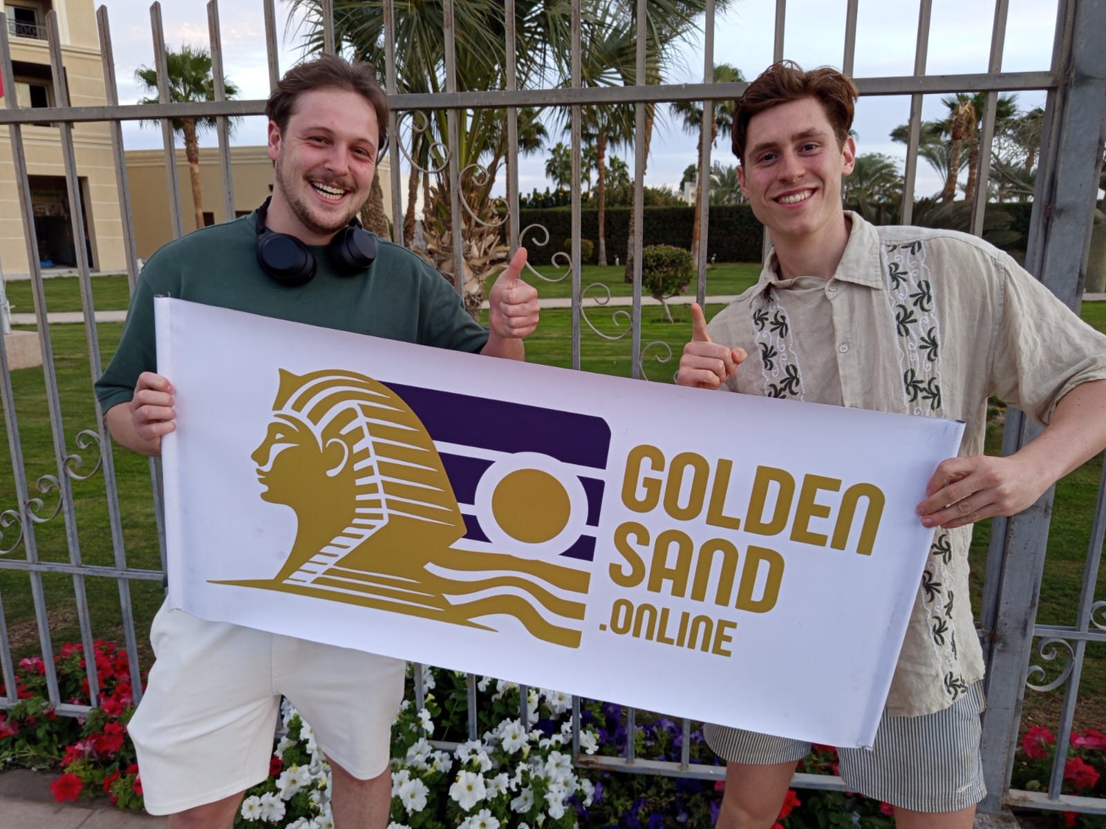 ضيفان مبتسمان يرفعان إبهاميهما ويحملان لافتة «Golden Sand» في حديقة نخيل خلف سياج حديدي