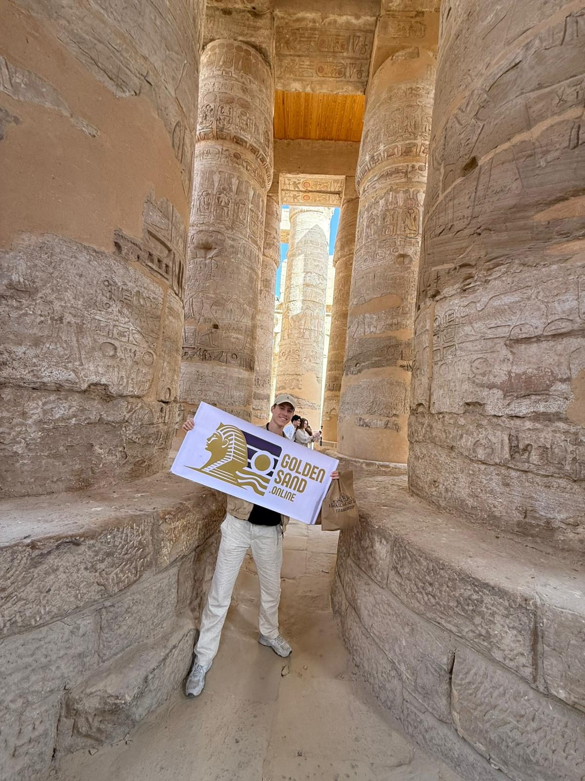 Guest holding the Golden Sand banner between giant hieroglyph-carved columns at Karnak Temple in Luxor