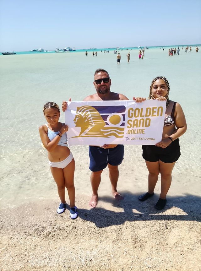 Three guests with a Golden Sand banner at the water's edge of a turquoise shallow bay, boats and waders behind