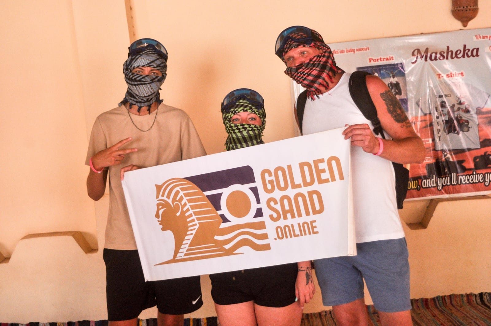 Three guests in desert scarves and goggles posing with a Golden Sand banner at a safari camp