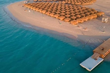 Children and adults on a sandy Red Sea beach with palm-thatched umbrellas beside clear turquoise water