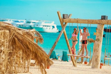 Couple on a wooden beach swing with thatched huts and white boats on turquoise water at Nemo Bay Island