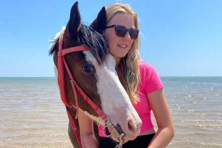 Smiling woman in sunglasses standing beside a brown and white horse with a red bridle at the Red Sea shore