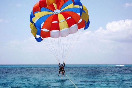 Two parasailers hanging below a red, yellow and blue parachute high above deep blue sea