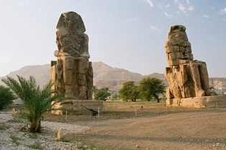 Two seated Colossi of Memnon on a dusty plain with a palm tree and desert hills behind near Luxor