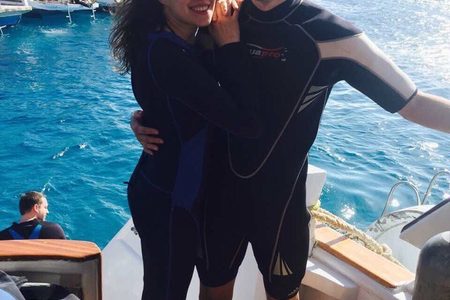 Smiling couple in dark wetsuits posing on the deck of a dive boat with other boats and blue sea behind them