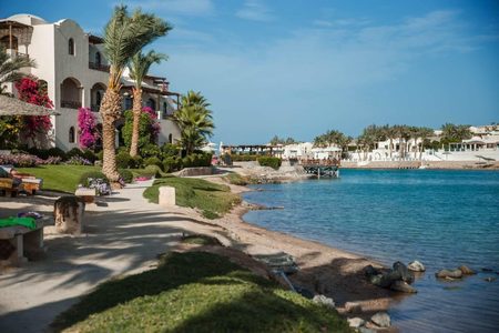 Whitewashed waterfront buildings with pink bougainvillea, palms and a wooden jetty beside calm blue water in El Gouna