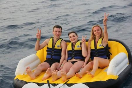 Three guests in life jackets on an inflatable sofa tube at sea, making peace signs