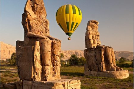 Yellow and green hot air balloon floating between the two Colossi of Memnon in golden light near Luxor