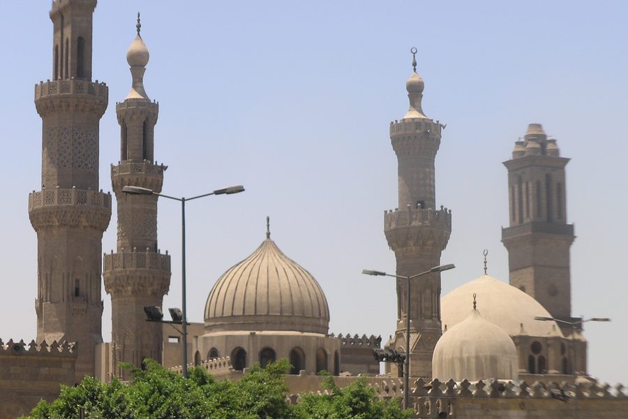 Minarets and domes of a historic mosque rising above trees in Islamic Cairo under a hazy sky