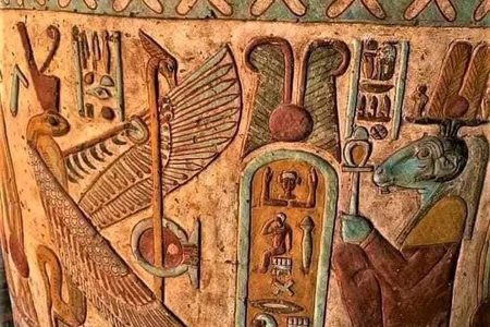 Colorful painted relief on a temple column showing a royal cartouche, a winged bird figure and a ram-headed god