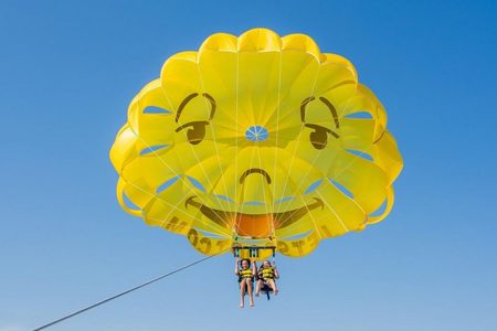 Two riders in yellow life vests parasailing under a yellow smiley-face parachute against a clear blue sky
