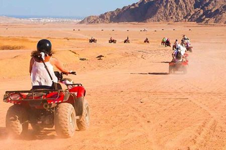 Rider in a black helmet on a red quad bike ahead of a line of quads crossing the desert