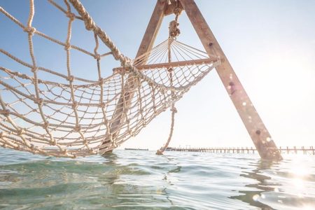 Rope net hammock hanging from a wooden frame over shallow sea at Orange Bay, with a long jetty behind