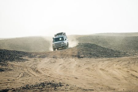 Safari jeep driving along a sandy desert track with a trail of dust under a hazy sky