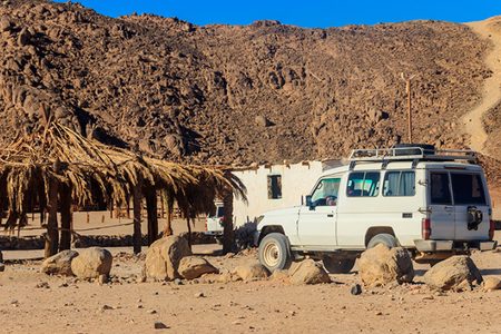 Safari jeep parked beside a palm-thatched shelter and a small white building at the foot of a rocky desert mountain