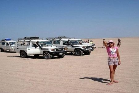 Tourist in a pink hat poses beside a row of white 4x4 safari jeeps on a flat sandy plain near Marsa Alam