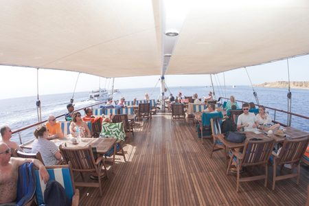 Tourists seated at wooden tables under a shade awning on the teak deck of a sailing boat at sea