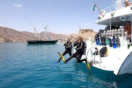 Two scuba divers in black wetsuits jumping off the back of a dive boat into turquoise water