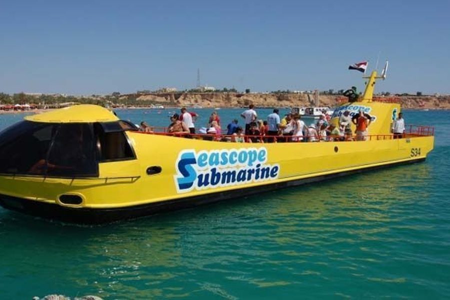 Passengers on the deck of a yellow Seascope Submarine boat cruising past a beach with umbrellas and low cliffs