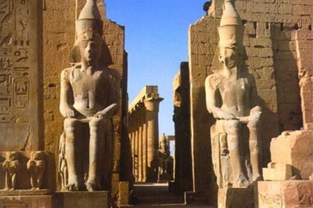 Two seated colossal pharaoh statues flank a temple gateway in Luxor, with columns visible beyond