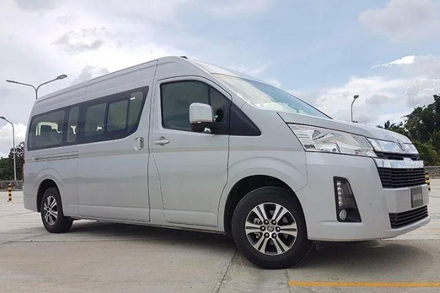 Un minibús Toyota Hiace plateado aparcado en un aparcamiento pavimentado con árboles y un cielo nublado al fondo
