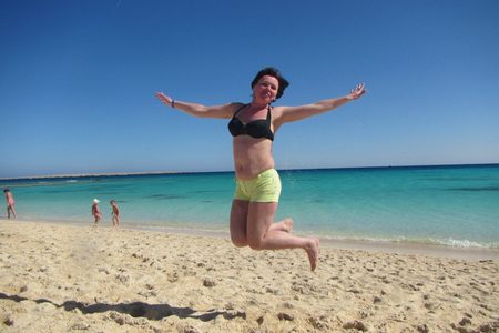 Tourist jumping with arms outstretched on a sandy beach beside turquoise sea while children play at the shore