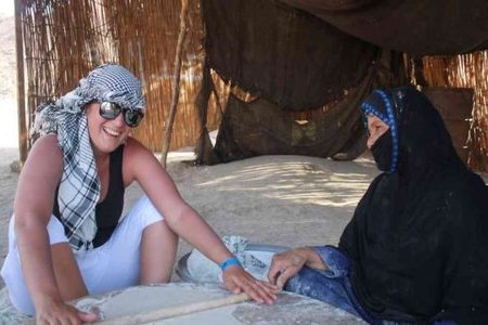 Tourist in a checkered scarf rolls dough with a veiled local woman inside a reed hut at a desert camp near Marsa Alam