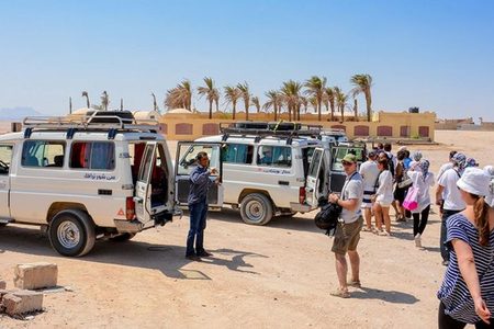 Tourists in sun hats and scarves listen to a guide beside two white safari jeeps at a desert stop