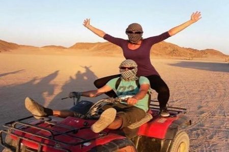 Two riders in face scarves on a red quad bike in warm evening light, one standing with arms spread wide