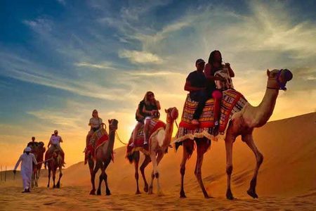 Tourists riding camels in a line across golden desert dunes at sunset, led by a guide in a white robe
