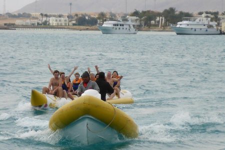 Tourists waving from a yellow banana boat towed by a speedboat, with white boats and palm trees on the shore