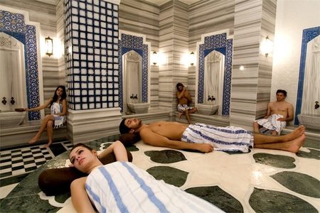 Guests wrapped in striped towels relaxing in a marble Turkish hammam with blue and white tiled niches