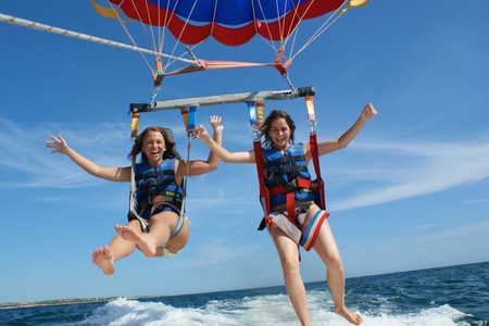 Two smiling women with arms outstretched parasailing in blue life vests above the sea and boat wake