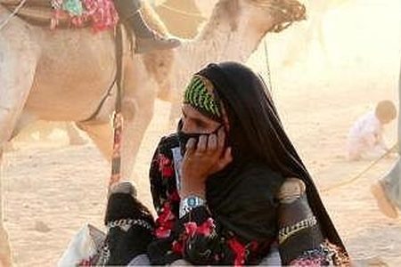 Veiled woman in a floral dress sits on desert sand beside a camel near Hurghada, with a man in a long robe behind her
