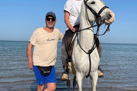 A rider on a white horse and a man standing beside it at the water's edge of a pebbly Red Sea beach in Hurghada