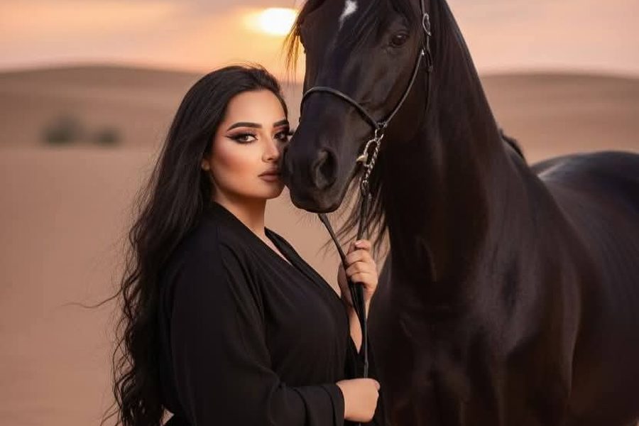 Woman in a black abaya standing beside a black horse on desert dunes at sunset