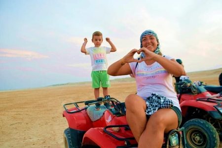 Woman making a heart with her hands beside a boy flexing his arms on red quad bikes in the desert near Hurghada