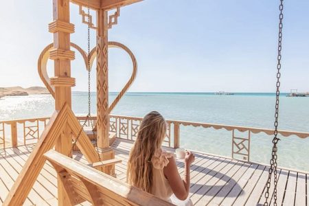 Woman with a cup on a wooden swing with a heart-shaped frame, looking over the turquoise sea at Orange Bay