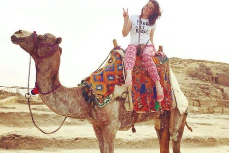 Woman riding a camel with a colorful saddle blanket, making a peace sign in front of an ancient stone pyramid