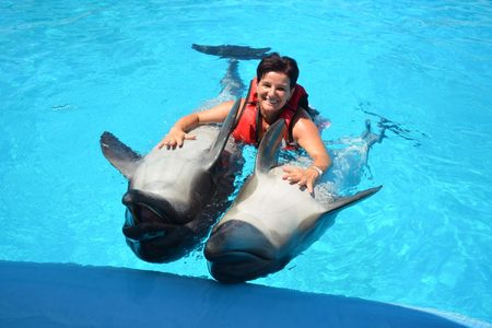 Smiling woman in a red life vest holding on to two dolphins as they swim through a bright turquoise pool
