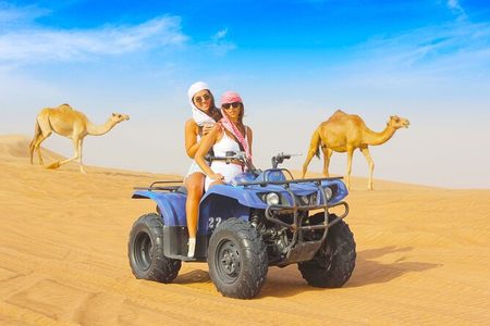 Two women in headscarves on a blue quad bike with camels walking across the desert behind