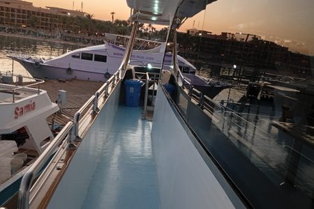 Side deck of the yacht in warm golden light with teak and chrome railings, docked boats and beachfront hotels behind